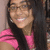 A person with long dark hair and glasses wears a pink shirt, smiling broadly in three showing slight head movement