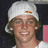 A person wearing a white cap with red and yellow details, smiling broadly against a dark background with pink elements.