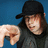 A person wearing a black cap and dark shirt points downward with their right hand against a blue background.