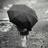 Black-and-white photo of a person holding an umbrella in rain, with red pixelated text on black background reading "I've been Waiting Since February For 75 Today Is The Day ...finally. Knowing it Won't come But I can't Help but Prey".