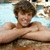 A shirtless person with brown hair lies on a light blue towel, arms crossed over chest in a pool setting.