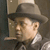 Static pixelated image of a man in profile wearing a fedora and dark suit with light shirt collar, set against neutral background.