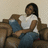 A pixelated image shows a person with dark hair wearing a white shirt and blue pants seated on a brown couch against a light beige background.