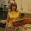 A woman in a yellow sleeveless top and patterned skirt stands in a kitchen holding a brown tray, with cabinets and appliances visible behind her.