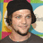 Pixelated image of a person with dark hair and a black beanie, wearing a dark shirt and black choker, against a colorful striped background with white cloud shapes.
