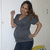 A woman in a gray top and dark pants poses with one hand on her hip and the other raised, standing against a light background with a yellow object partially visible.