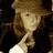 Pixelated image of a person with long brown hair wearing a wide-brimmed hat and light-colored clothing against dark background.