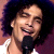 Pixelated static image of a person with dark curly hair holding a microphone, wearing a white sleeveless top against a purple background.