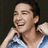 Pixelated close-up of a person with short dark hair wearing a light blue sleeveless top, hand near face in a relaxed pose.
