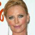 A pixelated close-up of a person with light brown hair styled in an updo, wearing a white top against a light background with partial orange text on the left.