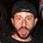 Pixelated close-up of a person wearing a dark cap and dark shirt, with visible facial features including eyes, nose, mouth, and beard against a dark background.