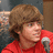 Pixelated image of a person with light brown hair wearing red clothing holding a silver microphone against a dark background.