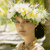 A pixelated figure with a large white and yellow floral headpiece, green foliage, and a black earpiece against a blurred green and yellow background.