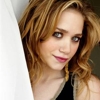 A close-up portrait of a person with wavy light brown hair, wearing a black sleeveless top and red lipstick, positioned next to a white vertical surface.