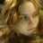 A pixelated close-up of a person's face with light skin tone, brown hair, and a neutral expression, set against a blurred background of warm brown and yellow tones.
