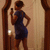 A woman in a blue dress stands before a mirror holding a phone, with one hand on her hip and the other near her face.