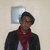 Pixelated image of a person wearing dark jacket, red scarf, and light shirt, standing against light wall with square mirror behind.