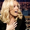 A static image of a person with blonde hair wearing a dark jacket, hand near chest, against a blurred cityscape background.