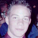 Static image of a person with short light-colored hair wearing dark blue clothing against a blurred background.