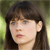 A static pixelated image of a person with long dark brown hair and bangs, light skin, and a neutral expression against a blurred green background.