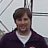 A pixelated static image shows a person with brown hair wearing a dark purple jacket over a white shirt against a light background with diagonal lines.