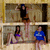 Animated sequence showing two women in a room with stone walls; one woman in a black top and brown shorts stands with arms raised while the other in a blue shirt sits holding a purple object.