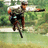 A person in a cap and light shirt rides a bicycle on a grassy path with trees, captured across showing motion