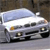 Pixelated image of a white sedan with dark windows and orange headlights, positioned on a gray road against a brown background.