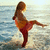 A woman in a bikini stands on a beach with one leg raised, facing the ocean under a bright sky.
