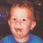 Pixelated close-up of a child's face with light skin, short brown hair, and blue clothing against dark background.