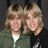 Pixelated static image of two people with light brown hair and neutral expressions, wearing dark tops against a dark background.
