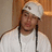 A pixelated image of a person wearing a white head covering and long dark hair, with a light-colored shirt against a brown background.