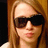 Pixelated close-up of a person with long light brown hair wearing dark sunglasses and a white top against a warm-toned background.