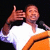 Pixelated image of a person in white and orange clothing making a hand gesture against black background.
