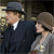 Two figures in period attire stand side-by-side; the left figure wears a dark suit and bowler hat, the right figure wears a light-colored dress and cloche hat.