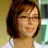 Pixelated image of a person with short brown hair wearing glasses and a white top against a blurred background.