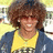 A pixelated image of a person with curly brown hair wearing sunglasses, a yellow shirt, and dark jacket against a blurred outdoor background.