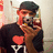 A person wearing a dark cap and black shirt with a red heart and white text, holding a camera to their face in a mirrored setting.