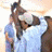 A horse and rider in a stable, with the rider adjusting gear while holding the reins.