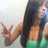 A person with long dark hair wearing a green top makes a peace sign gesture against a light background.