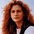 A pixelated close-up of a person with long, wavy reddish-brown hair wearing a black jacket over a white shirt against a light blue background.