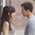 Two people face each other in a close-up view, one with long dark hair and the other with short dark hair, both in light-colored clothing against a blurred background.