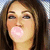 Pixelated close-up of a person with long brown hair blowing a large pink bubblegum bubble, mouth open, eyes partially closed, against a blurred background.