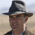 A pixelated close-up of a person wearing a dark wide-brimmed hat with a light band, standing against a blurred background of light brown and pale blue tones.