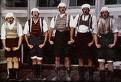 Static image of five men in traditional Bavarian-style clothing standing side-by-side against a building background.