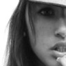 Black-and-white close-up of a person's face with long hair, wearing an eye patch and holding something near the mouth.