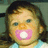 Pixelated close-up of a child's face with dark hair, light skin, and blue eyes holding a pink pacifier in their mouth.