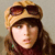 Pixelated image of a person wearing a light brown aviator cap with dark brown goggles resting on top, long brown hair, and a dark red garment against a neutral background.