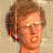 A pixelated image of a person with light brown hair and glasses, wearing a blue shirt, standing outdoors near greenery.