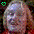 Pixelated static image of a stylized face with multicolored pixel blocks, featuring a green heart symbol in the top-left corner against a dark background.